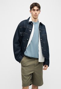 Νέος άνδρας που φοράει ένα σκούρο τζιν μπουφάν πάνω από ένα ανοιχτό μπλε μπλουζάκι και ένα λευκό πουκάμισο από μέσα, συνδυασμένο με χακί σορτς, στέκεται μπροστά σε λευκό φόντο.