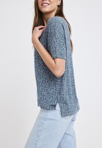 Kortärmad topp i lätt tyg i teal med ett blommönster i rosa och vitt, med rundad nederkant och sidospalter.