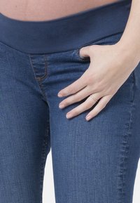 Jeans de maternidade azul escuro com uma cintura elástica, corte slim e detalhes de costura subtil; textura denim suave. Mão apoiada no quadril.