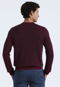 Mann mit lockigem Haar, der nach rechts schaut. Er trägt einen bordeauxfarbenen gemusterten Pullover und marineblaue Hosen und hat die Hände in den Taschen.