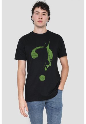 Junger Mann trägt ein schwarzes T-Shirt mit grünem Batman-Gesicht im Fragezeichen-Design, steht vor einem einfarbigen hellgrauen Hintergrund.