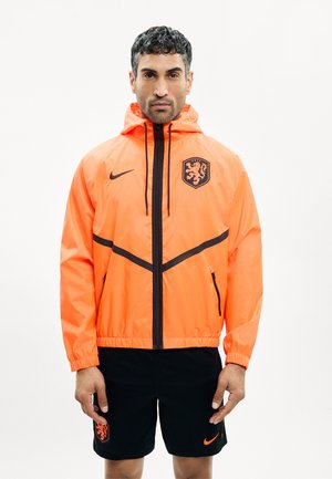 Mann trägt eine leuchtend orange Kapuzenjacke mit schwarzem Reißverschluss und Löwenemblem, kombiniert mit schwarzen Shorts mit passendem Emblem und Logo.