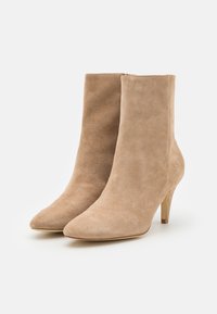 En par beige mockaskinnankelboots med spetsiga tår och smala klackar mot en vit bakgrund.