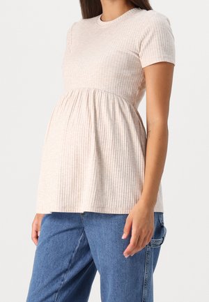 Gravid kvinde iført en ribstrikket, kortærmet beige graviditetstop kombineret med løstsiddende blå jeans mod en ensfarvet baggrund.