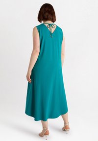 Turkos ärmlös klänning med en flödande silhuett, v-rygg och knytdetalj. Mjuk tyg och knälång nederdel, kombinerad med sandaler med remmar.