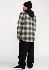 Kariertes Flanellhemd in grün, schwarz und weiß mit einem schwarzen Hoodie darunter; kombiniert mit lockeren schwarzen Hosen und schwarzen Sneakers.