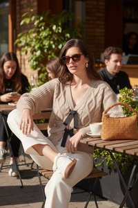 Mujer con gafas de sol y suéter beige está sentada con las piernas cruzadas en una mesa de café al aire libre con café y un bolso marrón claro.