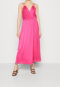 Robe maxi fuchsia à fines bretelles ajustables, col en V profond et taille froncée. Texture soyeuse tombant jusqu'aux chevilles, complétée par des talons noirs à lanières.