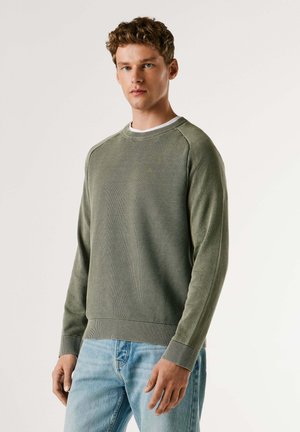 Zelený pletený sveter s rebrovanými manžetami a lemom, so zaobleným výstrihom a jemným vzorom. Nosený cez bielu košeľu, v kombinácii s modrými džínsami.