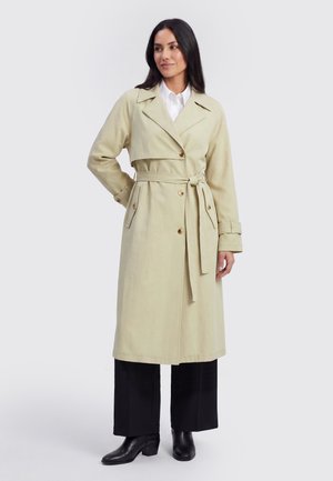 Donna con capelli lunghi e scuri che indossa un trench beige con cintura sopra pantaloni neri e stivaletti neri, in piedi con una mano sul fianco.