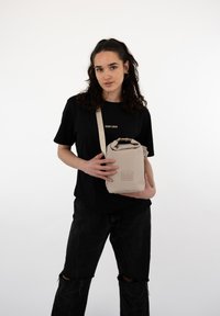 Borsa beige a scomparsa con finitura testurizzata, forma rettangolare e un'etichetta sul davanti, tenuta da una persona che indossa una maglietta nera e jeans scuri.