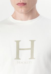 Hvit bomull t-skjorte med et stort lysebeige "H"-logo og ordet "Hackett" under i matchende farge, rund hals.