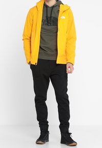 Chaqueta amarilla con capucha sobre un sudadera verde con un logo negro, combinada con joggers negros ajustados y calzado negro con suelas marrones.