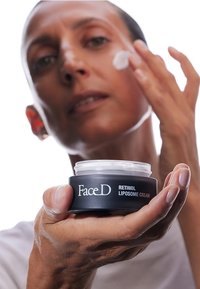 Barattolo nero etichettato "Crema Liposomiale al Retinolo FaceD", contenente una crema bianca, tenuto in mano. Utente che applica la crema sul viso. Texture liscia.