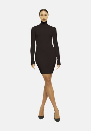 Donna in piedi con le gambe incrociate, indossa un abito nero attillato a maniche lunghe con collo alto e tacchi neri su uno sfondo semplice.