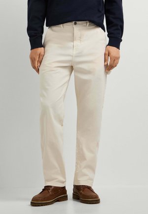 Mann trägt beige gerade geschnittene Hose, braune Leder-Schnürschuhe und ein dunkles Langarmshirt, steht vor einem einfarbigen Hintergrund.