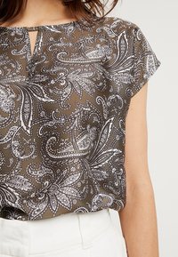 Durchsichtige braune Bluse mit schwarz-weißer Paisley-Musterung, rundem Halsausschnitt und kurzen Ärmeln. Schlüsselaugen-Detail an der Vorderseite.