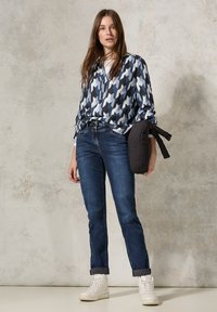 Blusa a maniche lunghe blu e bianca con un colletto, abbinata a jeans scuri e sneakers bianche alte, mentre tiene in mano un giubbotto di piumino nero.