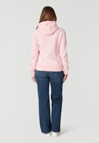 Sudadera con capucha rosa de corte relajado, con una capucha redondeada y costuras visibles. Combinada con unos jeans de pierna ancha en azul oscuro.