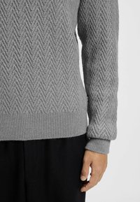 Grauer Strickpullover mit strukturiertem Fischgrätenmuster, mit gerippten Bündchen und Saum, kombiniert mit schwarzen elastischen Hosen.