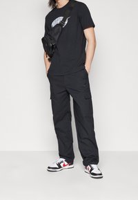 Man i svart grafisk t-shirt, svarta cargobyxor, vita sneakers med röd swoosh och svart crossbody-väska mot vit bakgrund.