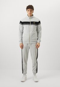 Lys grå joggedress med en zip-up jakke som har svarte striper og en logo, kombinert med matchende bukser som har svarte sidestriper og mansjetter.
