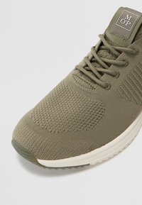 Oliwkowe buty sportowe z teksturowanej siateczki, minimalistyczny design i kontrastowa jasna podeszwa. Posiada oznaczenie marki na języku.