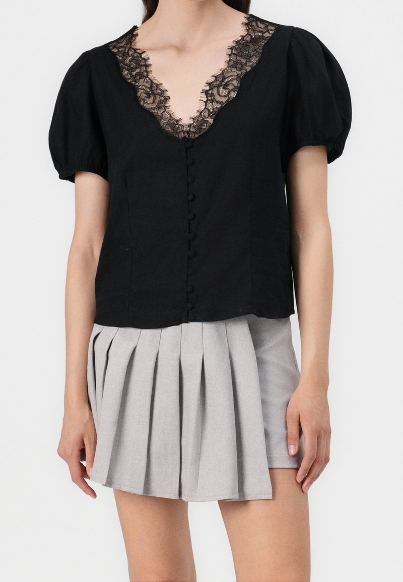 Blouse noire avec décolleté en V bordé de dentelle, manches bouffantes et fermeture à boutons sur le devant. Associée à une jupe plissée gris clair.