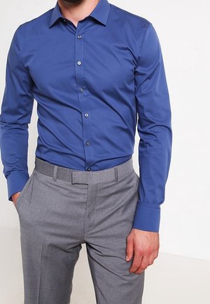 Homme portant une chemise bleue ajustée à manches longues rentrée dans un pantalon gris de costume, une main dans la poche et l’autre relâchée le long du corps.