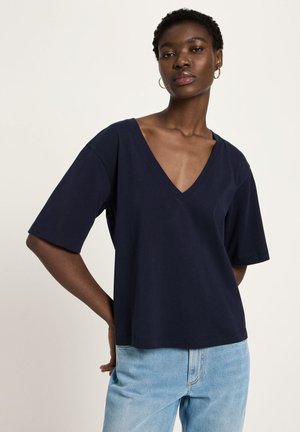 Woman with short curly hair wearing gold hoop earrings, a navy blue V-neck short-sleeve top, and light blue jeans stands with one hand behind back.