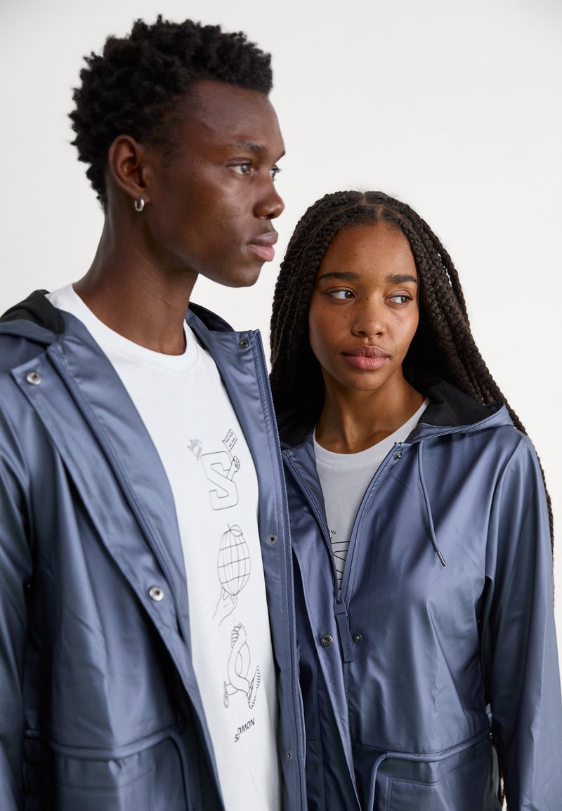 Grijze hooded jassen met een glanzende textuur, voorzien van ritssluitingen en zakken, gedragen over graphic T-shirts.