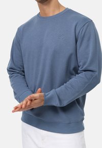 Homme portant un sweat à col rond à manches longues bleu avec un petit logo sur la poitrine, associé à un pantalon blanc, les mains jointes devant.