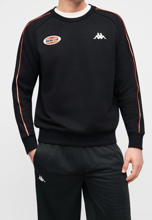 Homme portant un sweat-shirt noir avec des bandes orange sur les manches et un pantalon noir, tous deux avec des logos blancs Kappa. Le sweat-shirt possède un écusson Racing Club.