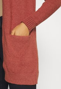 Rustkleurige cardigan van een zachte stof, met ribgebreide boorden en een voorzak. Eenvoudig, ontspannen ontwerp met een lange vorm.