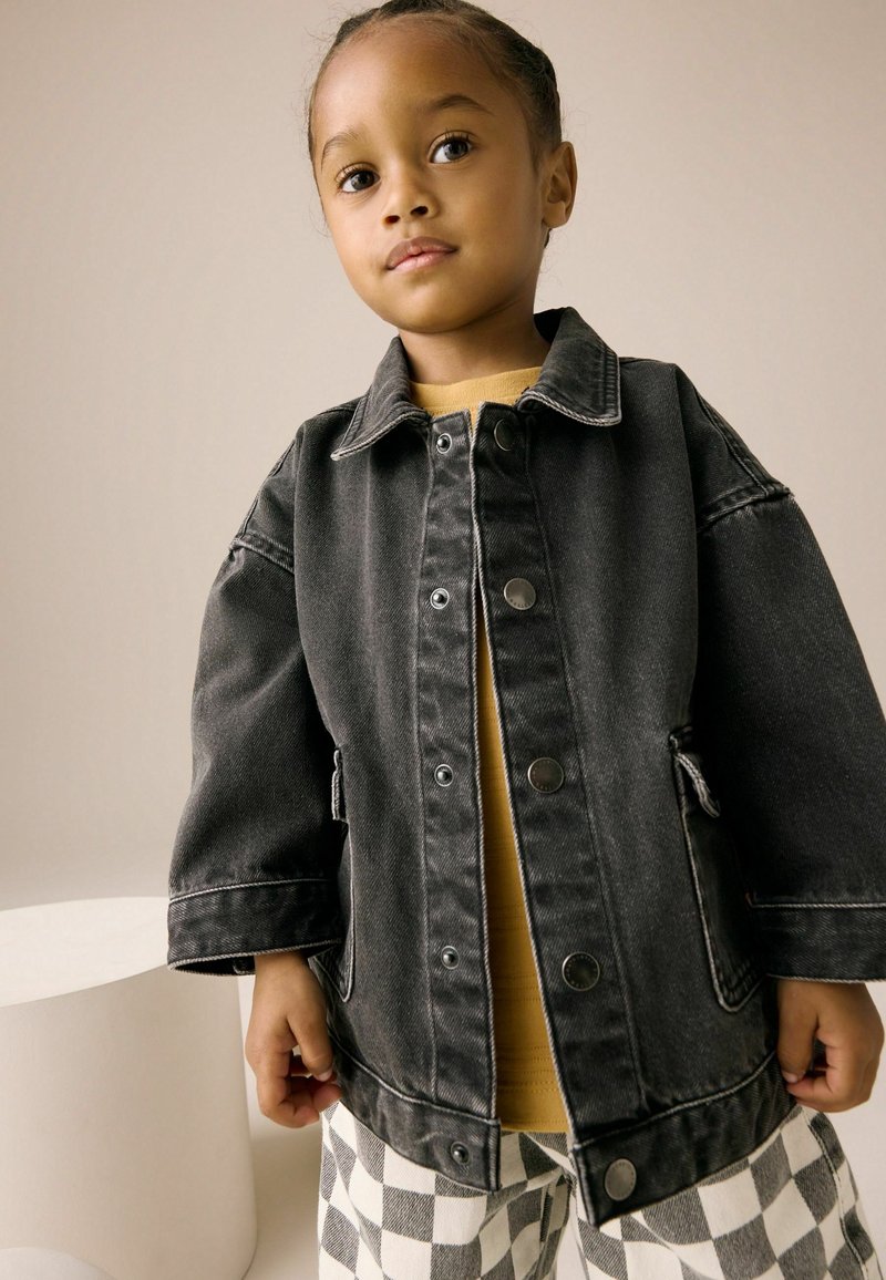 Enfant aux cheveux tressés portant une veste en jean foncée surdimensionnée, une chemise moutarde et un pantalon à carreaux noir et blanc, debout à l'intérieur.