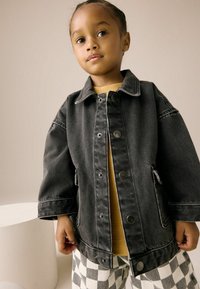 Veste en denim noir avec des boutons argentés, grandes poches et coupe décontractée, superposée à une chemise moutarde et un pantalon à carreaux.