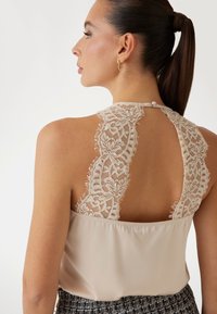 Bluză crem fără mâneci cu spate din dantelă, având modele intricate, guler rotund și închidere cu nasture la gât.