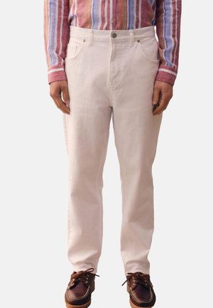 Hombre con pantalones beige claro, camisa de manga larga a rayas multicolores y zapatos marrón oscuro de cuero con cordones, de pie con las manos relajadas.