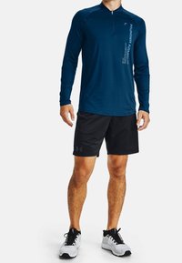 Long-sleeve navy athletic shirt with collar and half-zip. Black shorts and white athletic shoes. Under Armour logo on shorts.