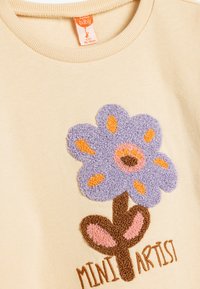 Sudadera de color crema con un diseño texturizado de flores moradas y acentos en naranja y rosa, y el texto "MINI ARTIST" en color marrón.