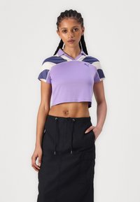 Polo roxo cropped com decote em V e mangas curtas, apresentando riscas diagonais em azul marinho e branco. Saia preta com bolsos tipo cargo.
