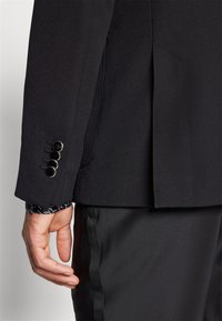 Jachetă neagră cu formă structurată, cu detalii la manșetă cu patru butoane și textură de țesătură netedă, asociată cu pantaloni negri.