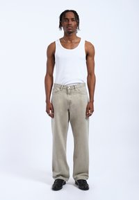 Hellgraue gerade Jeans mit einem verblassten Finish, kombiniert mit einem weißen Tanktop. Das Model steht aufrecht und trägt schwarze Schuhe.