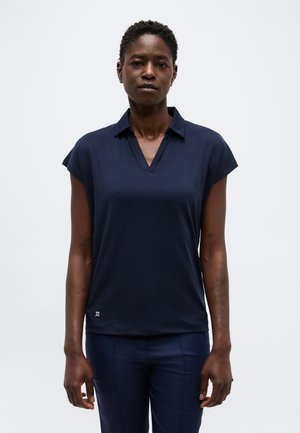 Sieviete ar īsiem matiem, ģērbusies tumši zila īsroku krekla ar apkakli un atbilstošas bikses, stāvot vienkrāsaina gaiša fona priekšā.