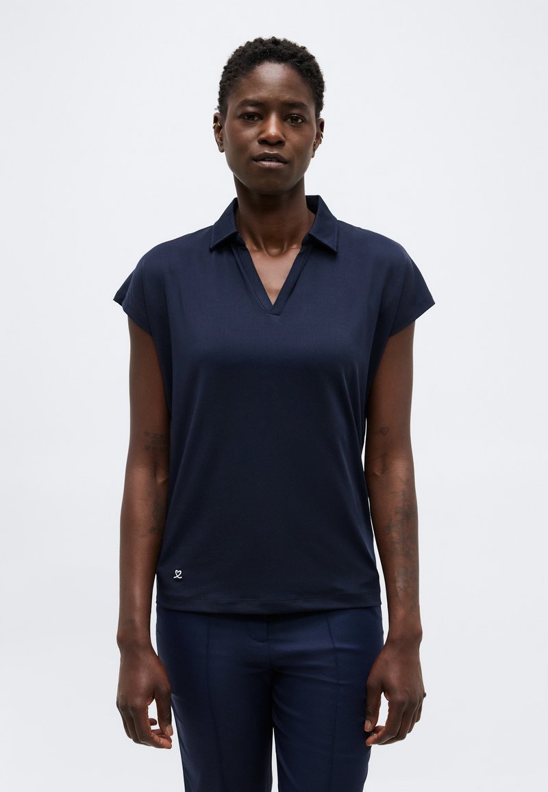 Γυναίκα με κοντά μαλλιά που φοράει ένα σκούρο μπλε κοντομάνικο πουκάμισο με γιακά και παντελόνι που ταιριάζει, στέκεται μπροστά σε ένα απλό φωτεινό φόντο.