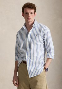 Jonge man met krullend haar die een wit-blauw gestreept overhemd met knopen, beige broek en een polshorloge met een bruine leren band draagt.