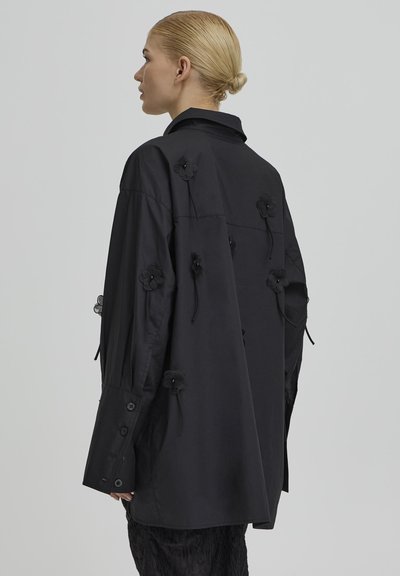Kvinde med blondt hår i en lav knold iført en oversized sort skjorte med sorte blomsterapplikationer på ryggen og ærmerne.
