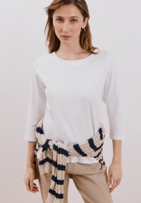 Maglietta bianca a maniche lunghe con dettagli in bottone dorato, abbinata a una sciarpa a costine beige e blu navy legata in vita, e pantaloni beige.