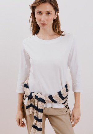 Weißes Langarmoberteil mit goldenen Knopfachsen, kombiniert mit einem beigen und marineblauen gestreiften Strickschal, der um die Taille gebunden ist, und beigen Hosen.