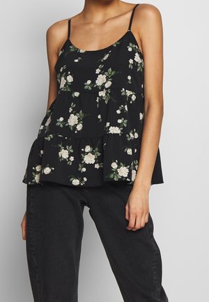 Femme portant un caraco noir à volants avec imprimé floral blanc et fines bretelles, associé à un jean noir, sur un fond uni.